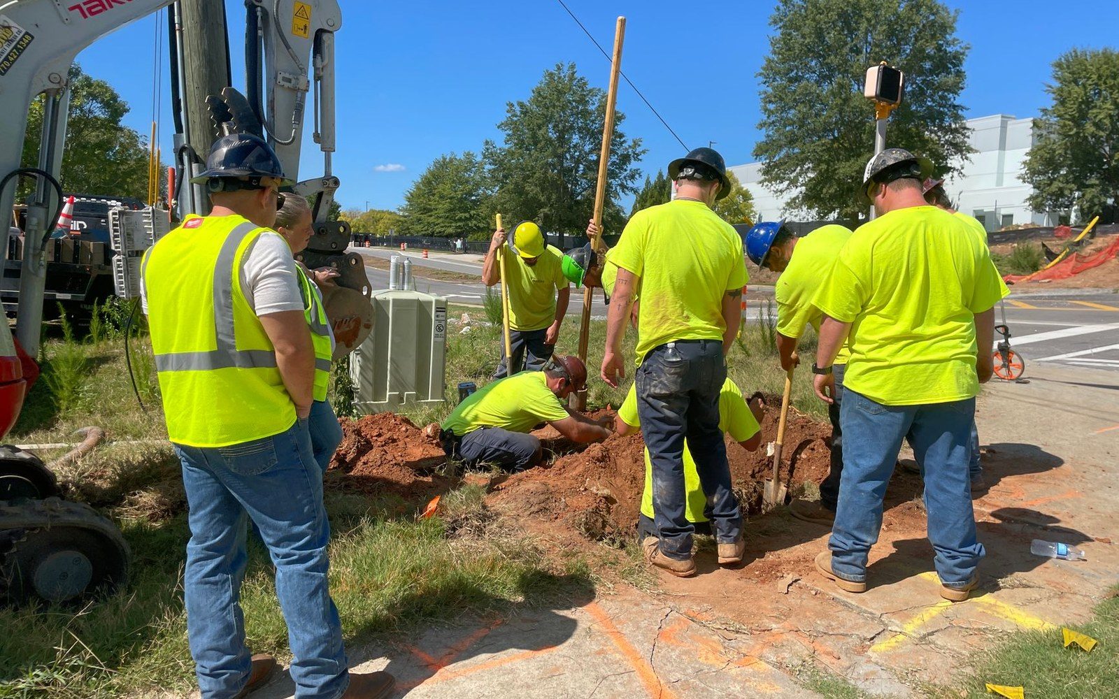 An open conduit trench cut along a parkway strip between sidewalk and kerb, a worker in hi-vis vest standing at its edge