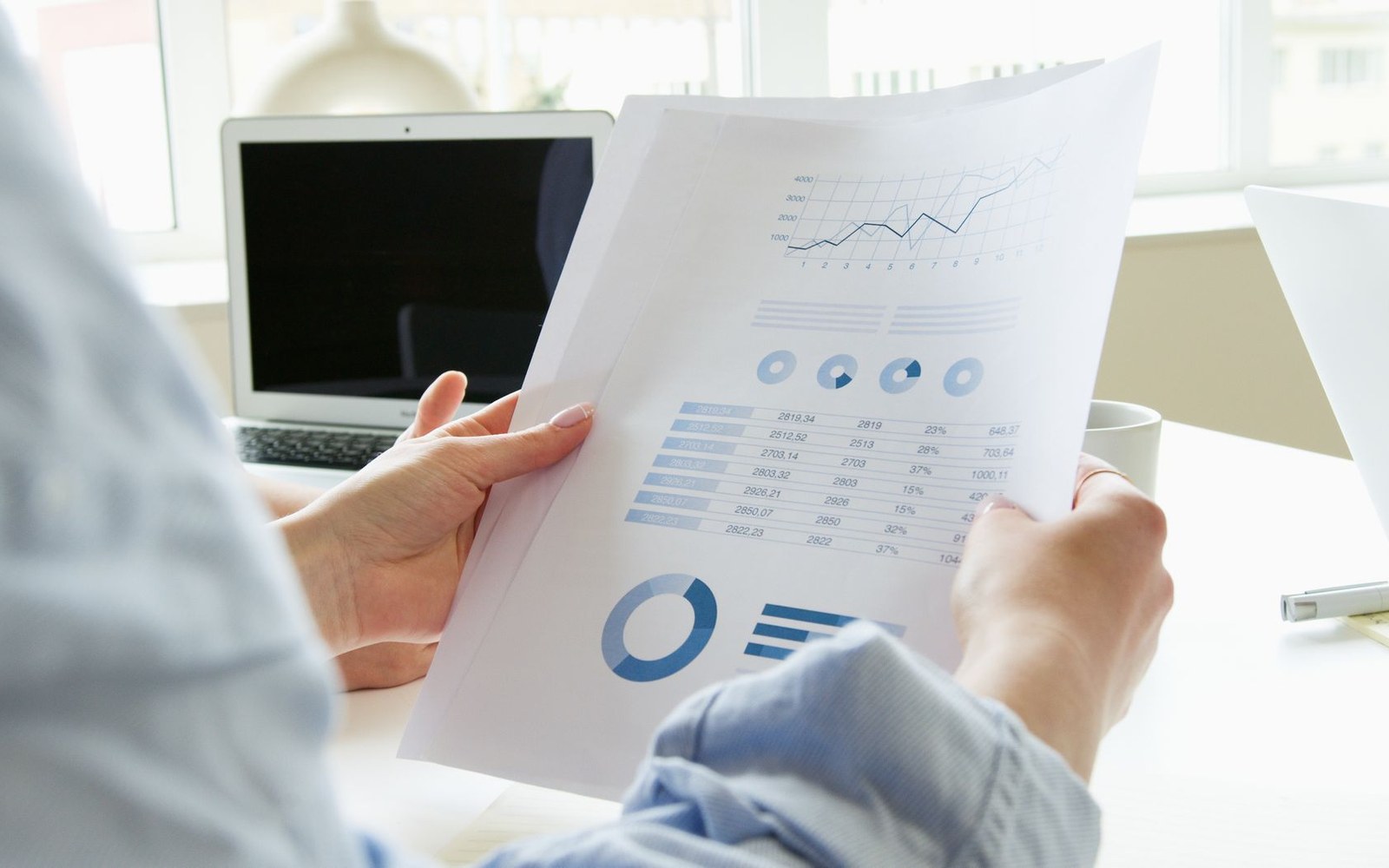 A printed broadband feasibility study open on a desk beside a laptop, a hand turning a page