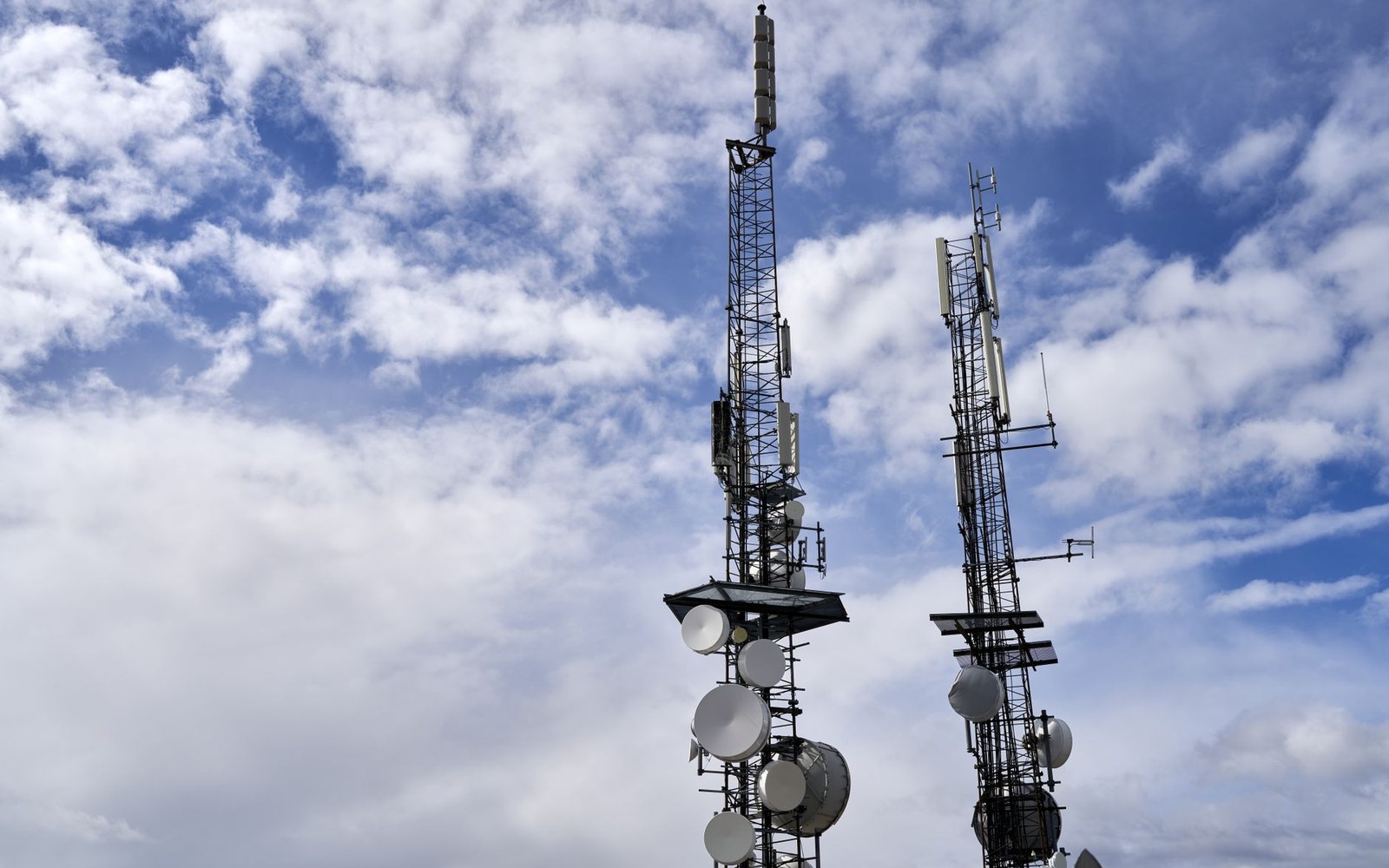 Two telecommunications towers with satellite dishes and antennas against a cloudy sky