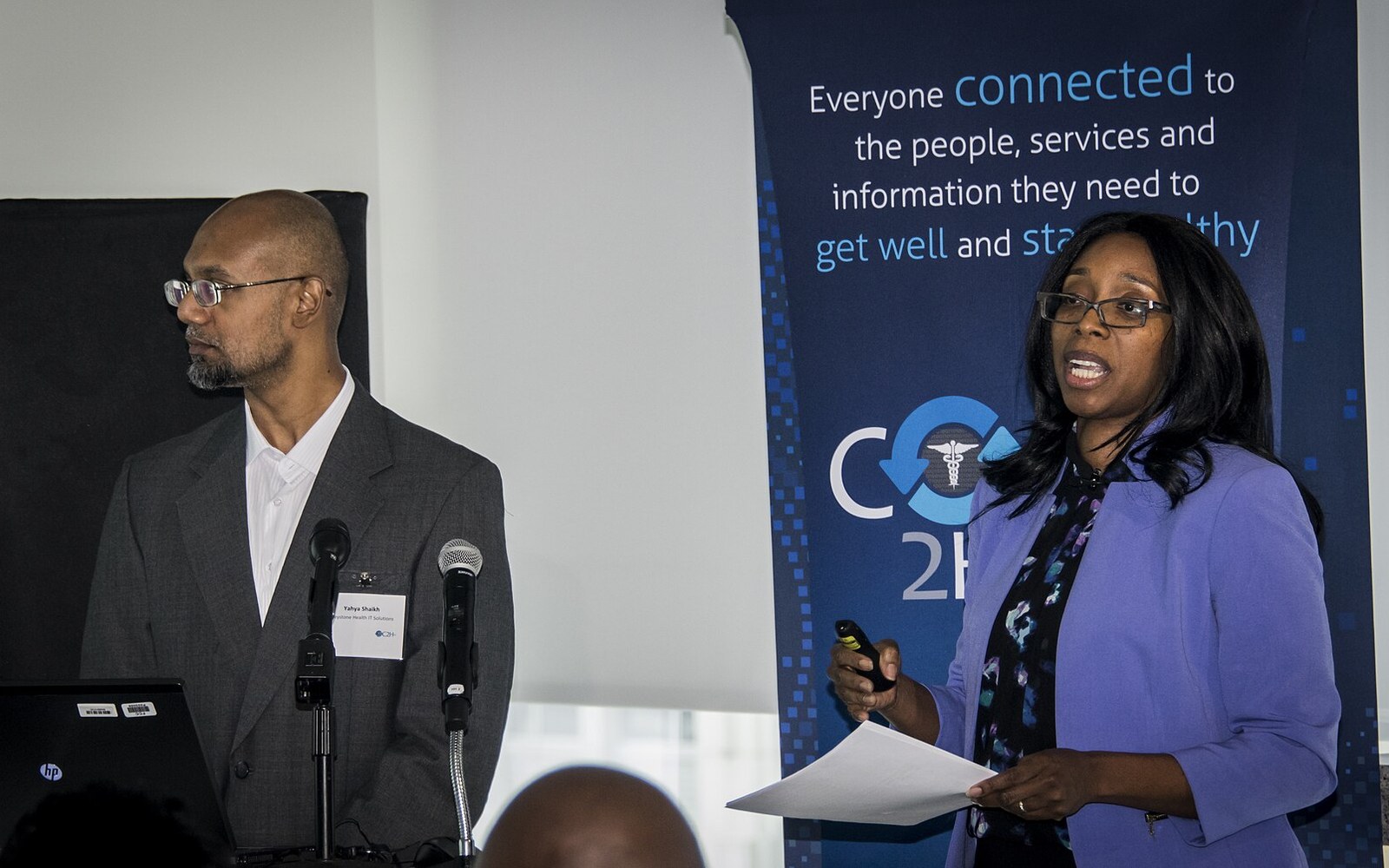 A woman in a purple blazer speaks at a podium beside a man near a conference banner