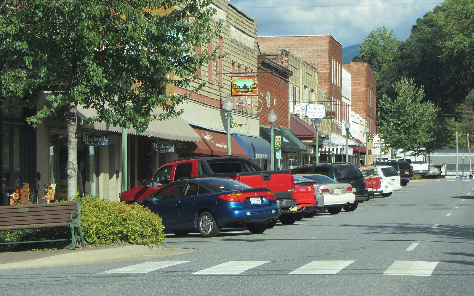 Cars parked along a small-town main street lined with brick storefronts and mountains beyond