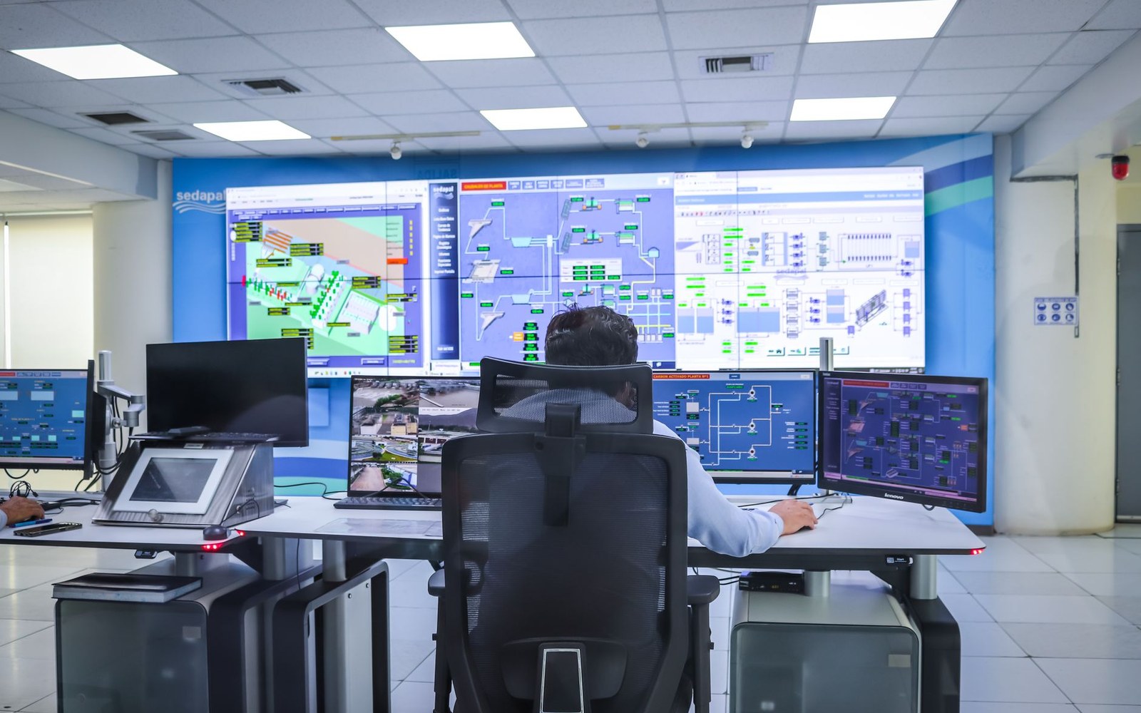 A network operations centre interior showing a wall of status monitors, an operator seated at a workstation in the foreground